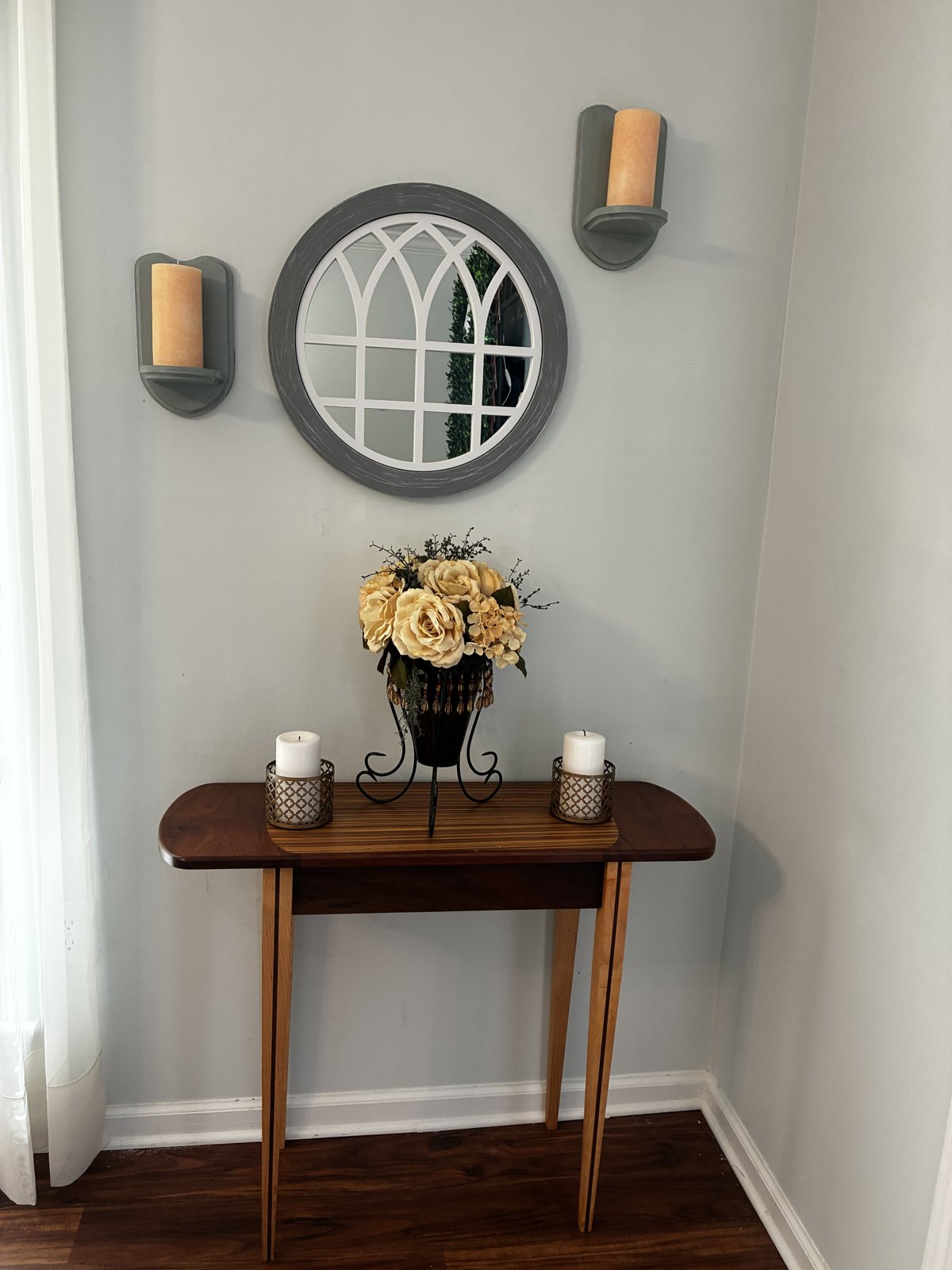 Wooden side table with a bouquet of ivory roses, two candles, a round arched mirror, and candle sconces on the wall